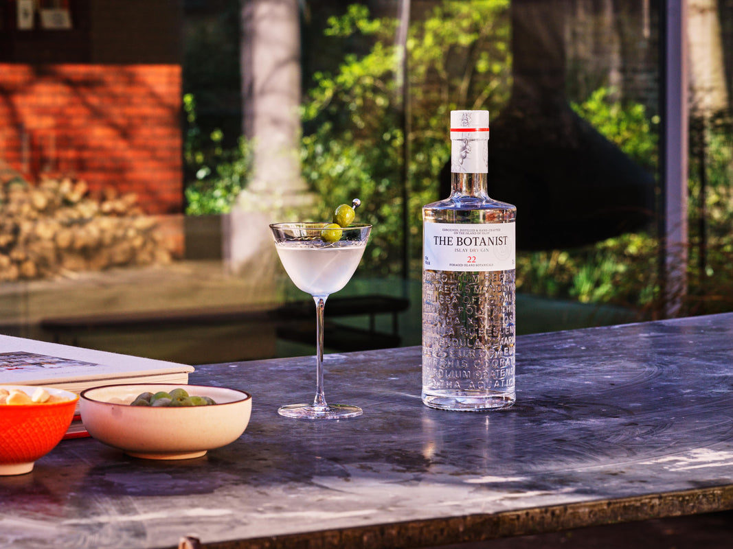 A lifestyle photo of a cocktail with olives, a Botanist bottle nearby, all on a counter top