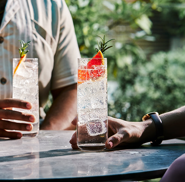 A lifestyle photo of two people holding fizzy cocktails with rosemary leaves